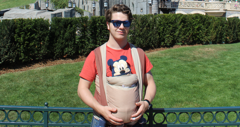 Ich mit Sonnenbrille vor einer Wiese mit Hecke. An meinem Bauch das Baby im Tragetuch, vollständig verdeckt. Es schaut nur der Kopf heraus, der mit einer Mickey-Maus-Mütze bedeckt ist.